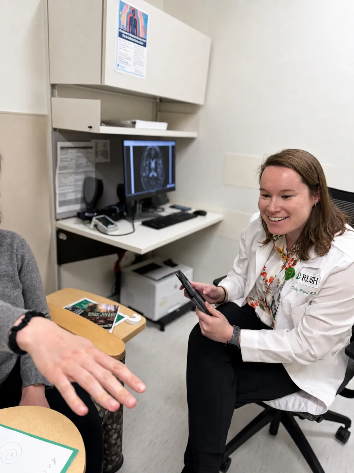 A neurologist with a patient during a movement disorders clinic visit, the patient's brain MRI on the screen behind them